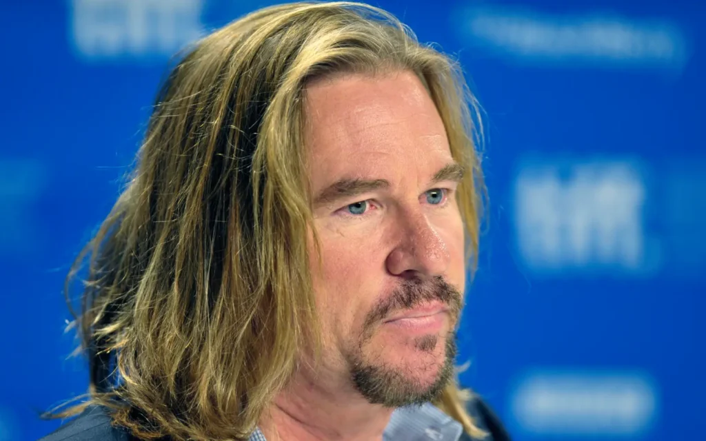 Val Kilmer, aktor Hollywood terkenal, berambut panjang dengan ekspresi serius di sebuah konferensi pers, berlatar belakang biru.