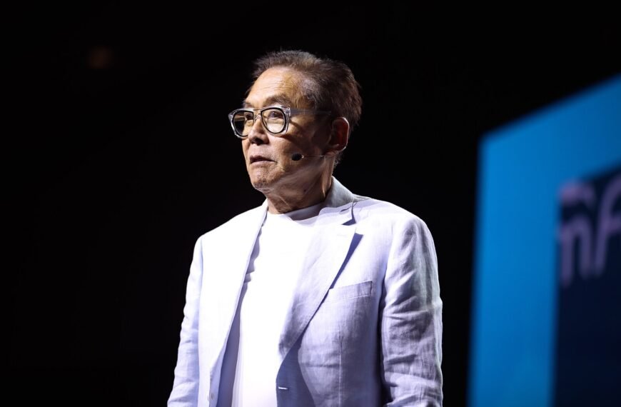 Robert Kiyosaki mengenakan jas putih saat memberikan pidato di atas panggung