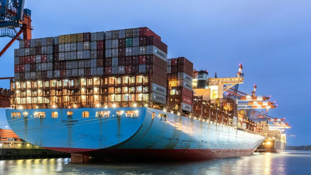 Sebuah kapal kargo besar yang penuh dengan kontainer berlabuh di pelabuhan pada malam hari, dengan derek pelabuhan yang aktif di latar belakang, melambangkan perdagangan internasional dan logistik ekspor-impor.