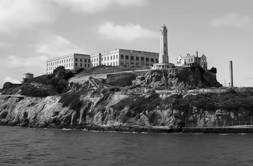Foto hitam putih Alcatraz pada era 1960-an, menampilkan bangunan utama penjara dan menara mercusuar di tepi Teluk San Francisco.